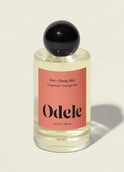 A clear bottle of Odele Hair + Body Mist, featuring grapefruit zest and orange peel scent, a black round cap, and a peach label, stands on a light surface with a soft shadow.