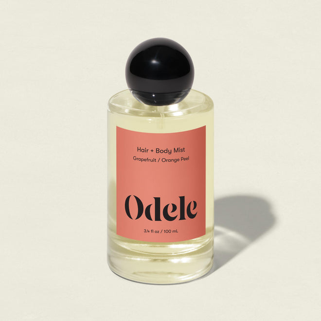 A clear bottle of Odele Hair + Body Mist, featuring grapefruit zest and orange peel scent, a black round cap, and a peach label, stands on a light surface with a soft shadow.