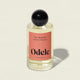 A clear bottle of Odele Hair + Body Mist, featuring grapefruit zest and orange peel scent, a black round cap, and a peach label, stands on a light surface with a soft shadow.
