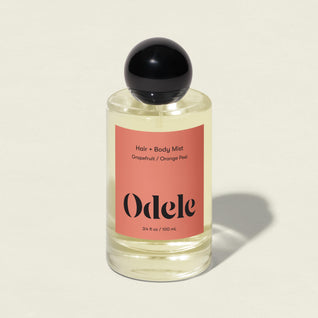 A clear bottle of Odele Hair + Body Mist, featuring grapefruit zest and orange peel scent, a black round cap, and a peach label, stands on a light surface with a soft shadow.