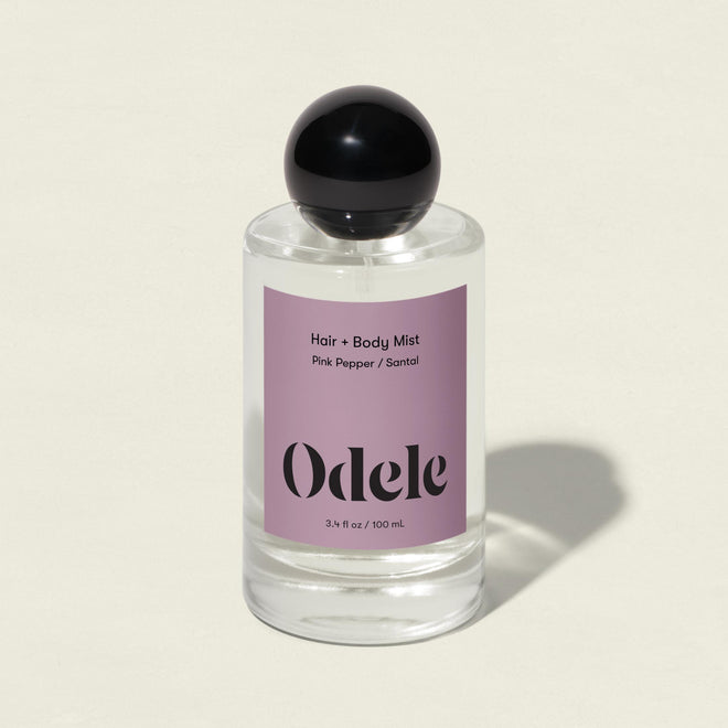 A clear glass bottle of Odele Hair + Body Mist with a black round cap and lavender label in the Pink Pepper / Santal scent, featuring pink peppercorn and sandalwood notes, sits against a light beige background.