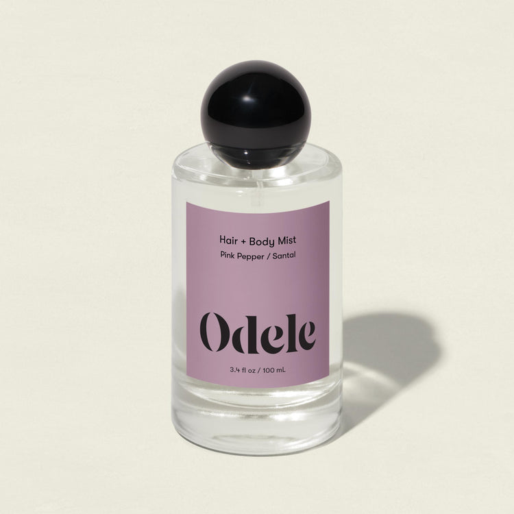 A clear glass bottle of Odele Hair + Body Mist with a black round cap and lavender label in the Pink Pepper / Santal scent, featuring pink peppercorn and sandalwood notes, sits against a light beige background.