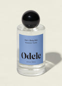 A clear glass bottle of Hair + Body Mist with a black round cap and blue label is shown on a light background. The label notes the vanilla, rosewood, and amber scent.