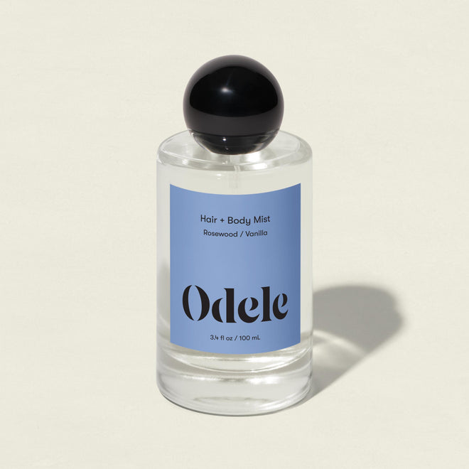 A clear glass bottle of Hair + Body Mist with a black round cap and blue label is shown on a light background. The label notes the vanilla, rosewood, and amber scent.