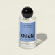 A clear glass bottle of Hair + Body Mist with a black round cap and blue label is shown on a light background. The label notes the vanilla, rosewood, and amber scent.