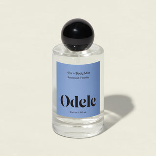 A clear glass bottle of Hair + Body Mist with a black round cap and blue label is shown on a light background. The label notes the vanilla, rosewood, and amber scent.