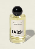 The Odele Signature Hair Scent, in a clear glass bottle with a black spherical cap and “Oakmoss / Ylang Ylang” label, is shown on a light beige background, suggesting fresh air with hints of cucumber and bamboo.