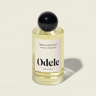 The Odele Signature Hair Scent, in a clear glass bottle with a black spherical cap and “Oakmoss / Ylang Ylang” label, is shown on a light beige background, suggesting fresh air with hints of cucumber and bamboo.