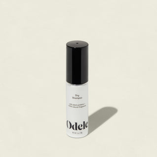 A white bottle of Dry Shampoo with a black cap stands upright on a light beige background, casting a soft shadow. Minimalist branding on the label highlights its oil-absorbing, volume-reviving formula.