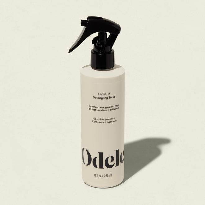 A white bottle of Odele Leave-in Detangling Tonic with a black spray nozzle stands upright on a light beige background. The label highlights plant proteins, natural fragrance, and heat protection for easy detangling.