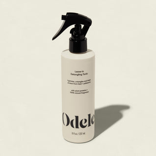 A white bottle of Odele Leave-in Detangling Tonic with a black spray nozzle stands upright on a light beige background. The label highlights plant proteins, natural fragrance, and heat protection for easy detangling.