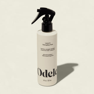 A white bottle of Odele Leave-in Detangling Tonic with a black spray nozzle stands upright on a light beige background. The label highlights plant proteins, natural fragrance, and heat protection for easy detangling.