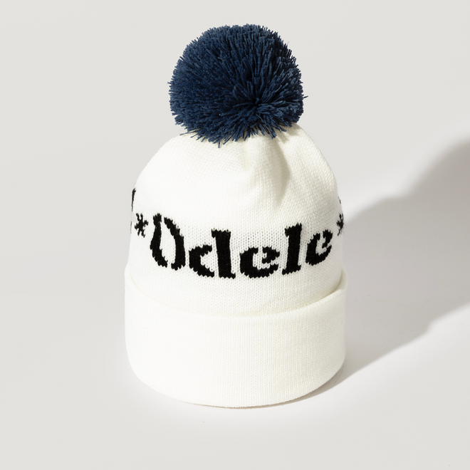 The Good Hair Day Winter Hat is a white knit beanie featuring a large navy blue pom-pom and bold black Odele lettering on the front—made by winter experts for quality comfort.