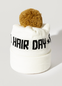 The Good Hair Day Winter Hat is a white knit beanie with a folded brim, a large brown pom-pom, and HAIR DAY boldly displayed in black on the front. Designed by winter experts for warmth and style.