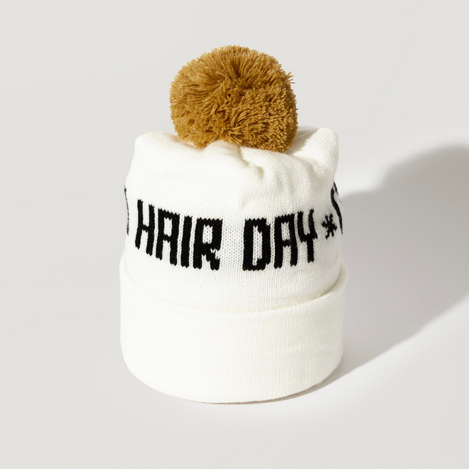 The Good Hair Day Winter Hat is a white knit beanie with a folded brim, a large brown pom-pom, and HAIR DAY boldly displayed in black on the front. Designed by winter experts for warmth and style.