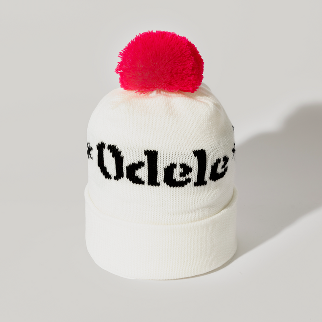 The Good Hair Day Winter Hat is a white knit beanie with a folded brim, bold black Odele text, and a large pink pom-pom—crafted by winter experts for comfort in this limited-edition style.