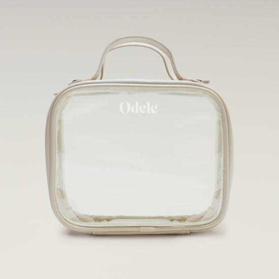 The Going Places Travel Bag is a clear, square-shaped essentials bag with beige trim and a top handle. Odele is printed in white on the front. Ideal for mini shampoos, it’s shown against a soft, neutral gradient background.