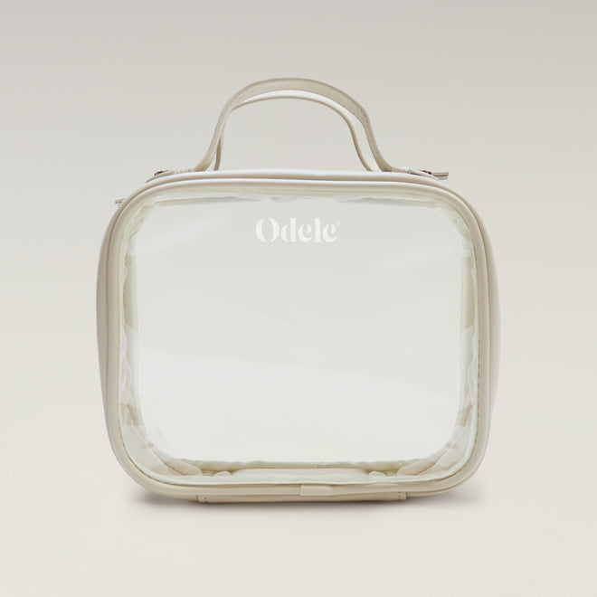 The Going Places Travel Bag is a clear, square-shaped essentials bag with beige trim and a top handle. Odele is printed in white on the front. Ideal for mini shampoos, it’s shown against a soft, neutral gradient background.