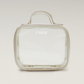 The Going Places Travel Bag is a clear, square-shaped essentials bag with beige trim and a top handle. Odele is printed in white on the front. Ideal for mini shampoos, it’s shown against a soft, neutral gradient background.