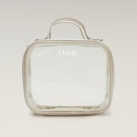 The Going Places Travel Bag is a clear, square-shaped essentials bag with beige trim and a top handle. Odele is printed in white on the front. Ideal for mini shampoos, it’s shown against a soft, neutral gradient background.