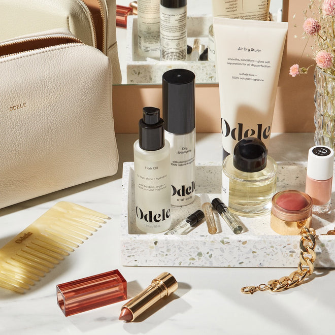 A variety of beauty products, including Odele salon-grade hair care, the Air Dry Styler Mini, lipstick, a comb, perfume vials, and a cream makeup pot, are displayed on a terrazzo tray with a beige cosmetic bag and gold bracelet.