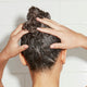 A woman with long dark hair applies Odele Scalp & Body Scrub to her hair in the shower.