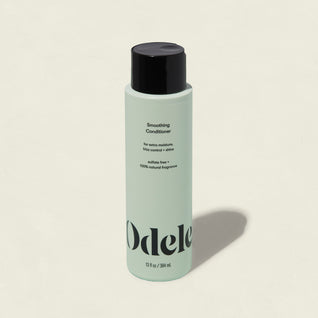 A light green bottle of Smoothing Conditioner with a black cap features text for extra moisture and frizz control. Sulfate-free with a natural fragrance, this conditioner enhances shine. The bottle is shown on a plain background.