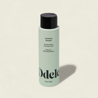 A light green bottle of Smoothing Shampoo with a black cap and minimalist branding on the front offers frizz control and shine, set against a plain, light background.