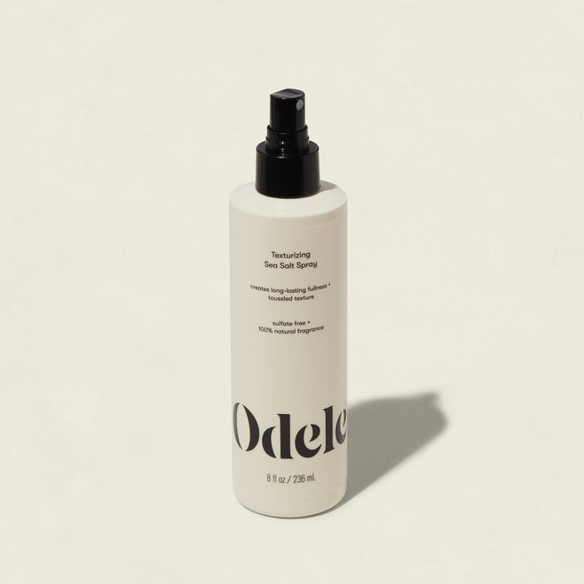 A white bottle of Odele Texturizing Sea Salt Spray with a black nozzle and minimalist label sits on a light background, casting a shadow to the right—ideal for creating effortless, tousled texture and that signature beach hair look.