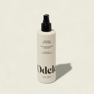 A white bottle of Odele Texturizing Sea Salt Spray with a black nozzle and minimalist label sits on a light background, casting a shadow to the right—ideal for creating effortless, tousled texture and that signature beach hair look.