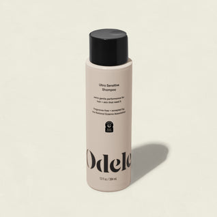 A beige bottle of Odele Ultra Sensitive Shampoo stands upright on a light background. The salon-grade shampoo, in black-capped packaging, displays sleek black text and the logo—ideal for sensitive skin and eczema-prone scalps.