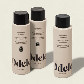 Three beige bottles from the Odele Ultra Sensitive Set—shampoo, conditioner, and body wash—stand upright on a light background. Their labels highlight that they are ultra sensitive and fragrance-free.