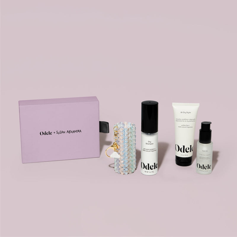 The Touch-Ups To Go Bundle is shown with a lavender Odele x Susan Alexandra gift box, beaded keychain, Hair Oil Mini, Air Dry Styler Mini, and a pump bottle of Odele hair care against a light pink background.
