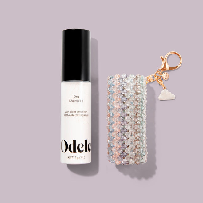 The Touch-Ups To Go Bundle includes a bottle of dry shampoo next to a pastel beaded keychain with a gold clasp and white cloud charm, displayed on a light purple background.