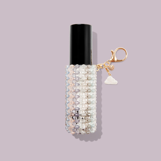 The Touch-Ups To Go Bundle features a cylindrical lip balm with a black cap in a beaded, transparent case and a gold keychain with a cloud charm—perfectly paired with your Hair Oil Mini—set against a pale lavender background.