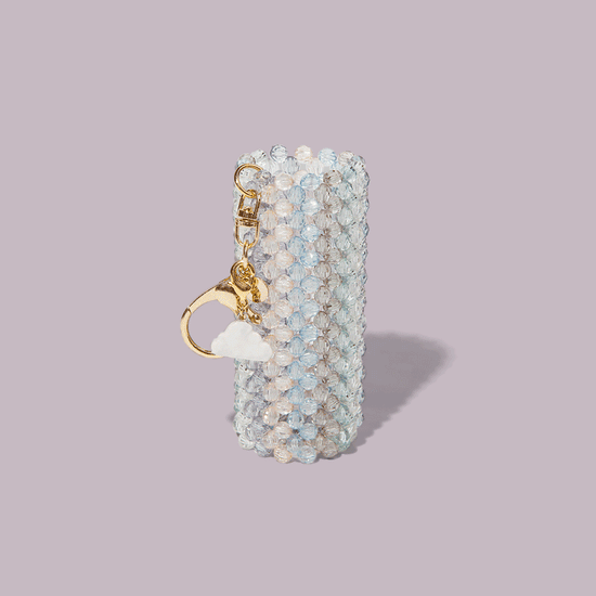 The Touch-Ups To Go Bundle features a cylindrical keychain holder with translucent beads, a gold keyring, cloud and bear charms—perfect for your Hair Oil Mini—shown on a light mauve background.