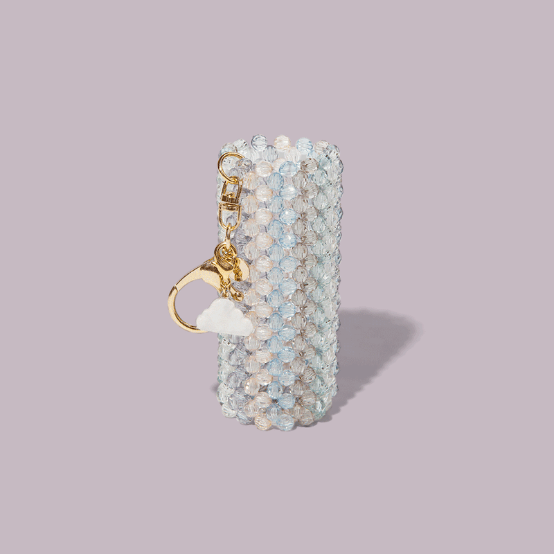 The Touch-Ups To Go Bundle features a cylindrical keychain holder with translucent beads, a gold keyring, cloud and bear charms—perfect for your Hair Oil Mini—shown on a light mauve background.