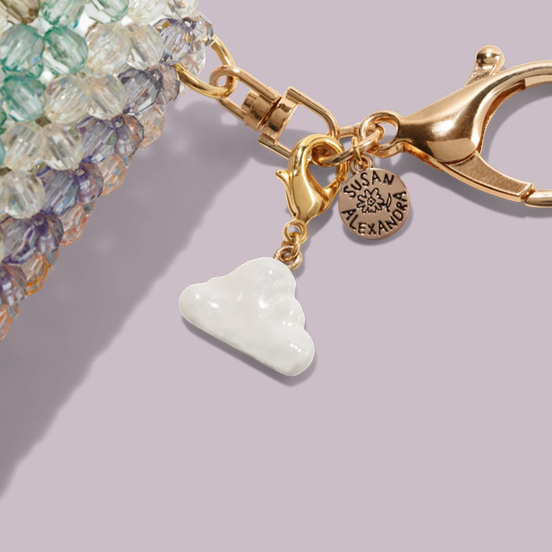A close-up of the Touch-Ups To Go Bundle: a beaded accessory with a gold clasp, Susan Alexandra engraved round charm, and white cloud-shaped charm—perfect next to your Hair Oil Mini—set on a light purple background.