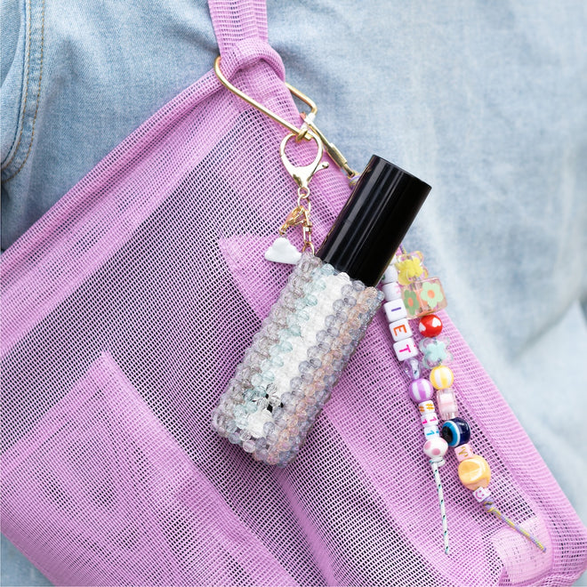 A person in a light blue denim jacket carries a pink mesh bag with the Touch-Ups To Go Bundle—a black mini hair oil in a beaded case—plus a colorful beaded keychain with letter and flower designs attached.