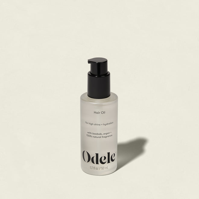A clear bottle of Odele Hair Oil with a black pump, labeled for high shine and hydration, rests on a light beige surface with its shadow extending to the right.