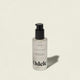 A clear bottle of Odele Hair Oil with a black pump, labeled for high shine and hydration, rests on a light beige surface with its shadow extending to the right.