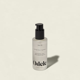 A clear bottle of Odele Hair Oil with a black pump, labeled for high shine and hydration, rests on a light beige surface with its shadow extending to the right.