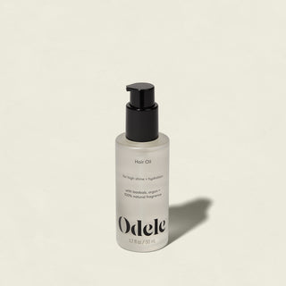 A clear bottle of Odele Hair Oil with a black pump, labeled for high shine and hydration, rests on a light beige surface with its shadow extending to the right.