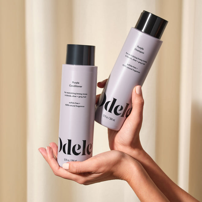 Two hands hold a bottle of Odele Purple Shampoo and Conditioner in front of a beige curtained background.