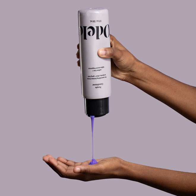 A person dispenses Odele Purple Conditioner onto their palm in front of a light purple background.