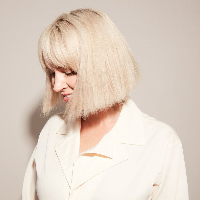 A woman with straight, chin-length platinum blonde hair smiles softly as she looks down, wearing an off-white collared shirt against a plain light background.