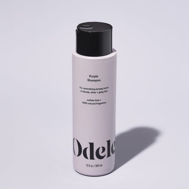 A bottle of Odele Purple Shampoo stands upright against a plain, light-colored backdrop with the front of the label facing the camera.