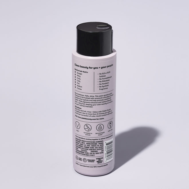 A bottle of Odele Purple Shampoo stands upright against a plain, light-colored backdrop with the back of the label facing the camera.