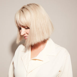 A woman with straight, platinum blonde hair in a blunt bob and bangs smiles softly. Her vibrant color, maintained with Odele Beauty Purple Shampoo to neutralize brassy tones, shines against her cream-colored blouse and a neutral background.