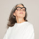 A woman with shoulder-length gray hair, kept bright and brass-free by Odele Beauty Purple Shampoo, wears black glasses and a white top. She tilts her head back and closes her eyes, appearing relaxed against a plain beige background.